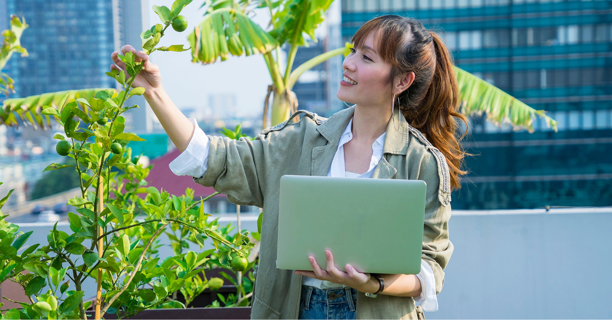 Woman examining plant while holding a laptop
